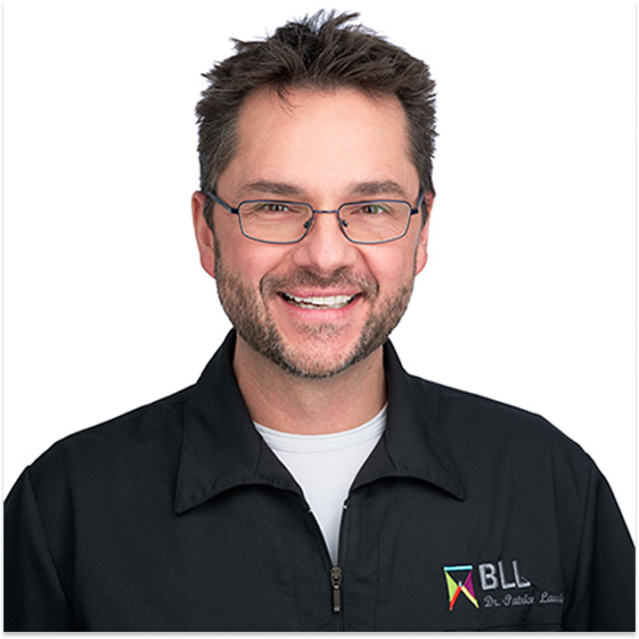 Dentiste souriant portant des lunettes, barbe courte et uniforme clinique.